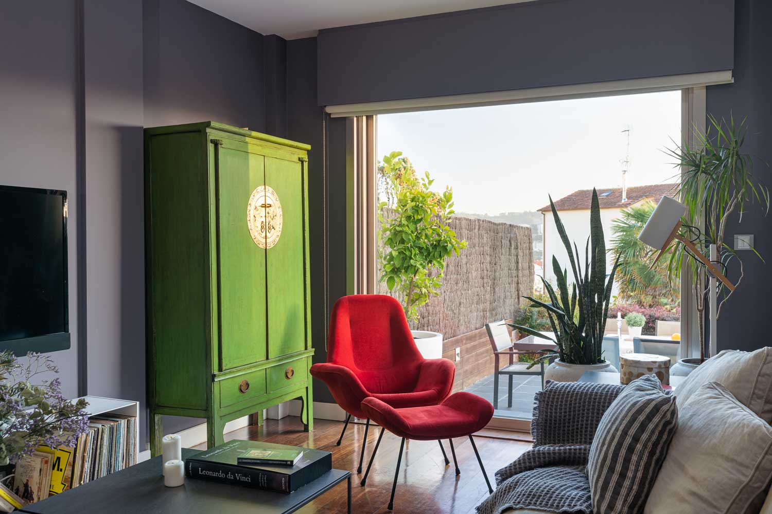 Detalle salón unifamiliar Oleiros_ sofá con textiles neutros y de rayas, butaca roja de lectura y armario de diseño verde junto a ventanal