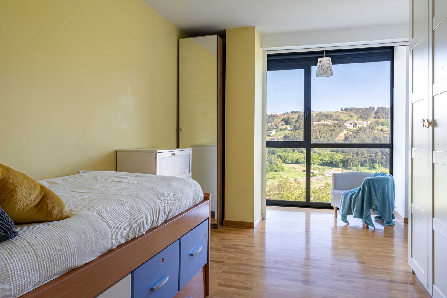 Dormitorio individual amarillo con gran ventanal y butaca junto a él_vistas a la naturaleza
