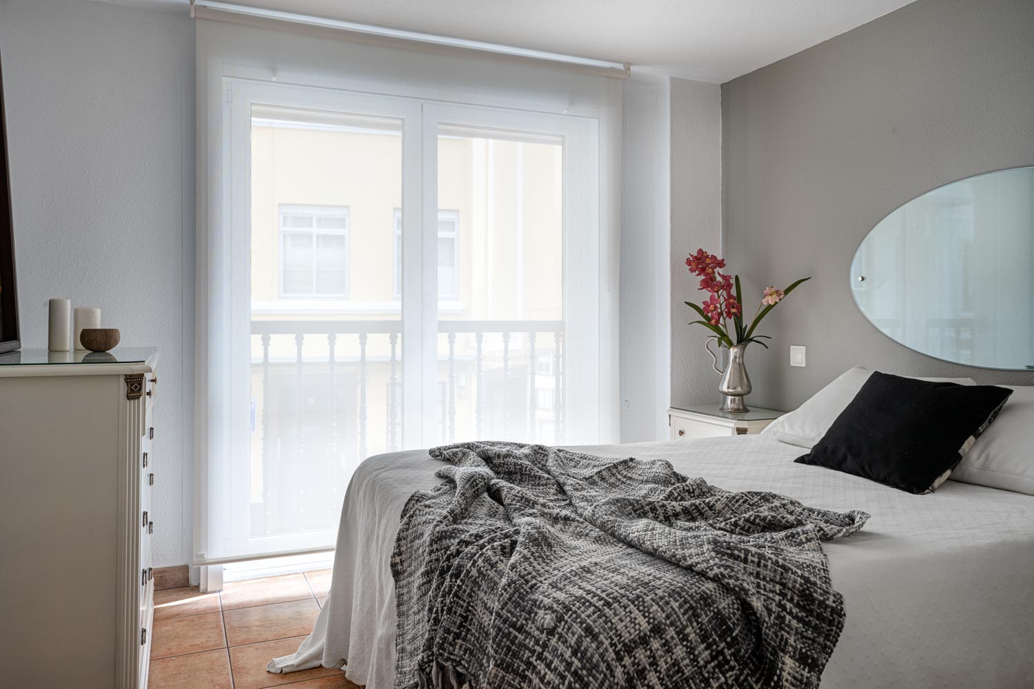 Dormitorio preparado con Home Staging con cama de matrimonio: Miramos hacia la ventana. Sobre la cama hay cojines y una manta y en la mesilla un jarrón plateado con flores
