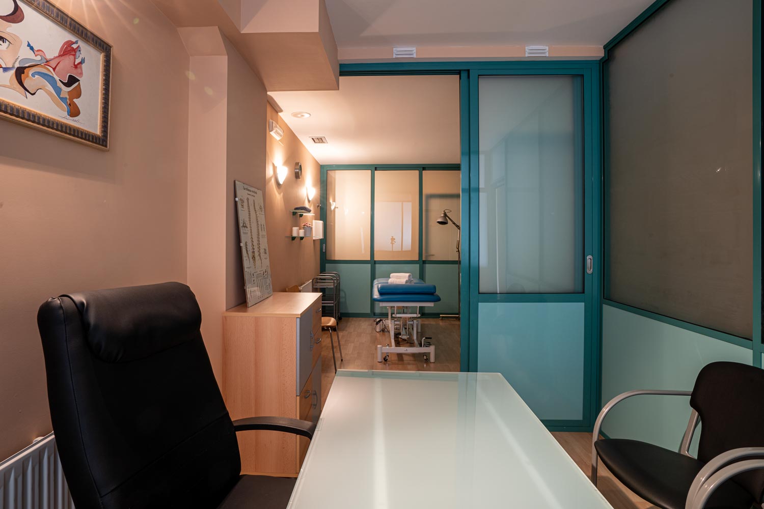 Interior de local comercial terapias naturales. Vemos el despacho y un gabinete con camilla azul comunicados por puertas verdes abiertas