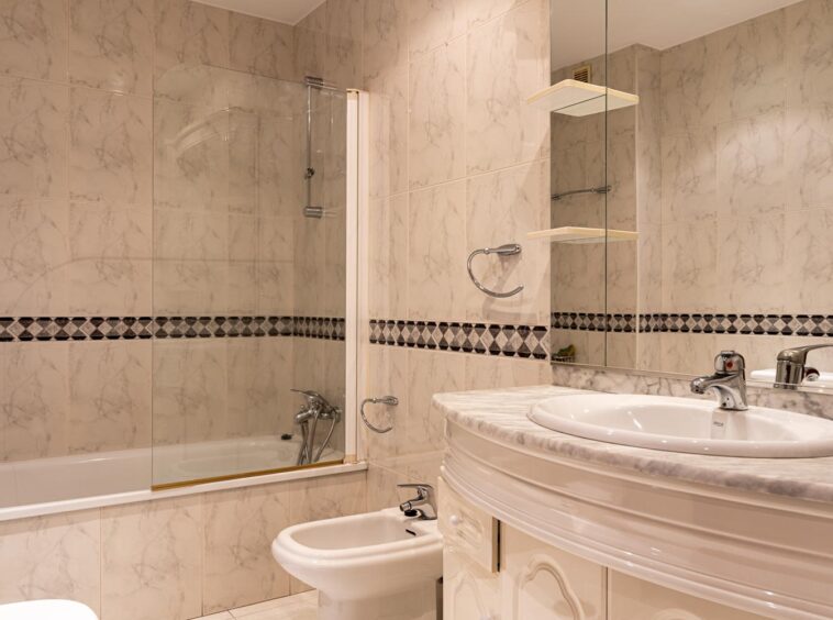 Cuarto de baño principal en piso de Betanzos_ Azulejo tipo mármol con cenefa. Se ve una bañera amplia, un bidé y un lavamanos con mueble redondeado y espejo