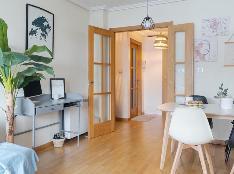 Salón-comedor luminoso y moderno con suelo de madera, una pequeña zona de escritorio y una mesa de comedor con sillas blancas y negras. La habitación está decorada con una planta grande, cuadros enmarcados y lámparas colgantes. Un pasillo con puertas de madera conduce a otras habitaciones.