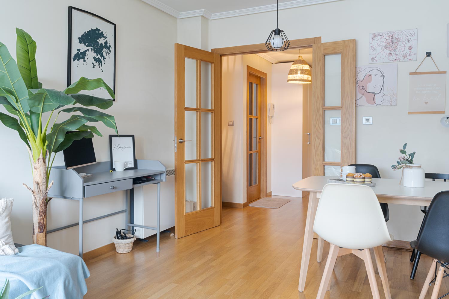 Salón-comedor luminoso y moderno con suelo de madera, una pequeña zona de escritorio y una mesa de comedor con sillas blancas y negras. La habitación está decorada con una planta grande, cuadros enmarcados y lámparas colgantes. Un pasillo con puertas de madera conduce a otras habitaciones.