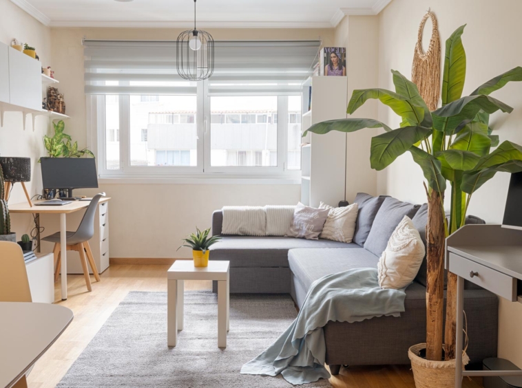 Salón luminoso con un gran sofá en forma de L, escritorio con ordenador, mesa de centro blanca y planta grande. La habitación tiene una ventana grande con persianas, estanterías con decoraciones y una lámpara colgante moderna.