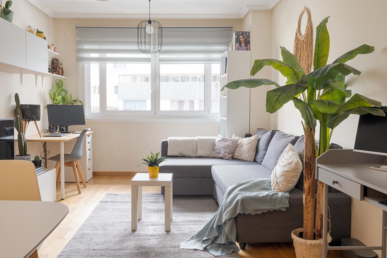 Salón luminoso con un gran sofá en forma de L, escritorio con ordenador, mesa de centro blanca y planta grande. La habitación tiene una ventana grande con persianas, estanterías con decoraciones y una lámpara colgante moderna.