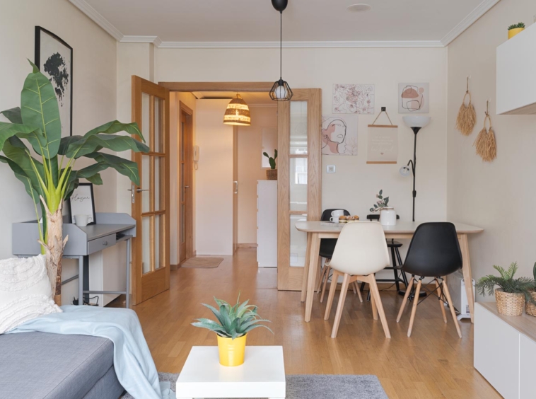 Sala de estar con decoración moderna, sofá gris, planta grande, escritorio pequeño y mesa de comedor con sillas blancas y negras. El pasillo con puertas de madera conduce a otras habitaciones. La habitación tiene paredes decoradas con cuadros y adornos colgantes.