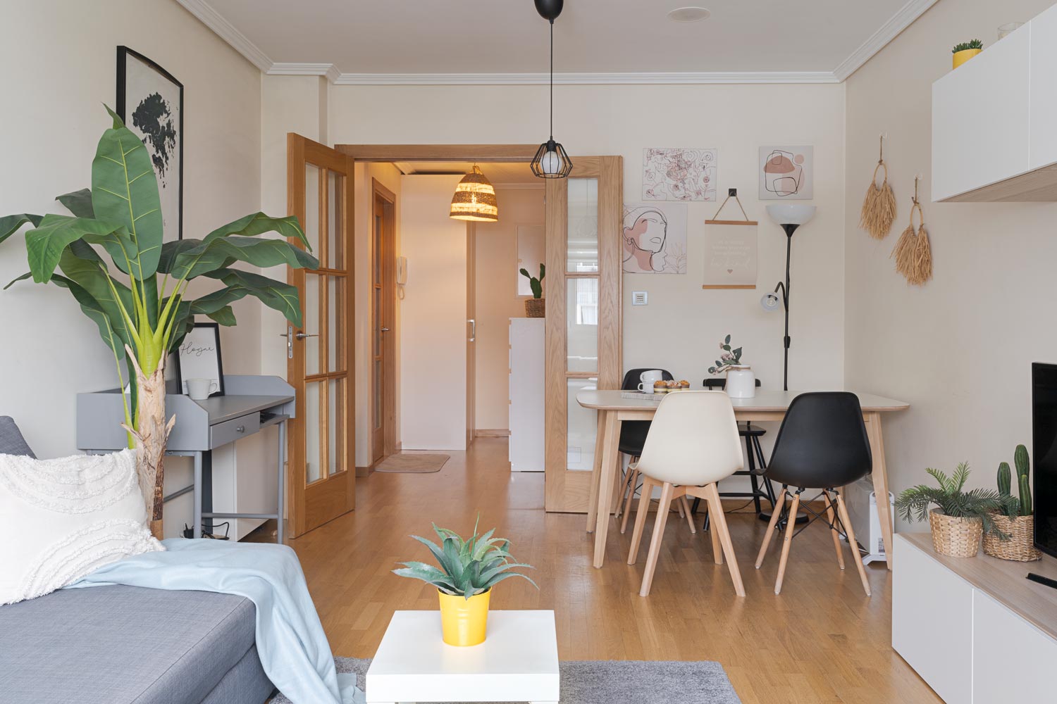 Sala de estar con decoración moderna, sofá gris, planta grande, escritorio pequeño y mesa de comedor con sillas blancas y negras. El pasillo con puertas de madera conduce a otras habitaciones. La habitación tiene paredes decoradas con cuadros y adornos colgantes.