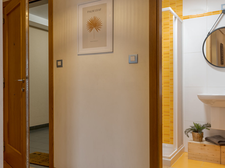 Pasillo con puerta de madera abierta que conduce a un baño con azulejos amarillos, lavabo blanco y espejo redondo. La pared del pasillo está decorada con un cuadro.