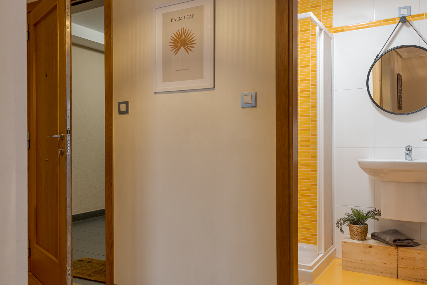 Pasillo con puerta de madera abierta que conduce a un baño con azulejos amarillos, lavabo blanco y espejo redondo. La pared del pasillo está decorada con un cuadro.