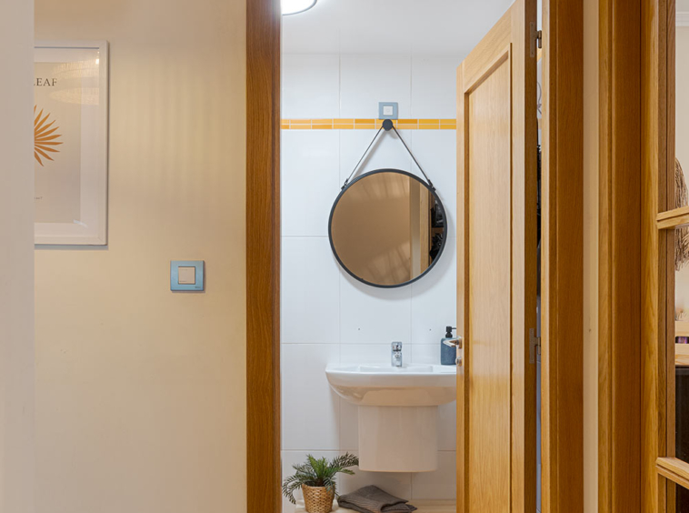 Vista desde el pasillo hacia un baño con azulejos amarillos, lavabo blanco y espejo redondo. La puerta de madera del baño está abierta, y la pared del pasillo tiene un cuadro decorativo.