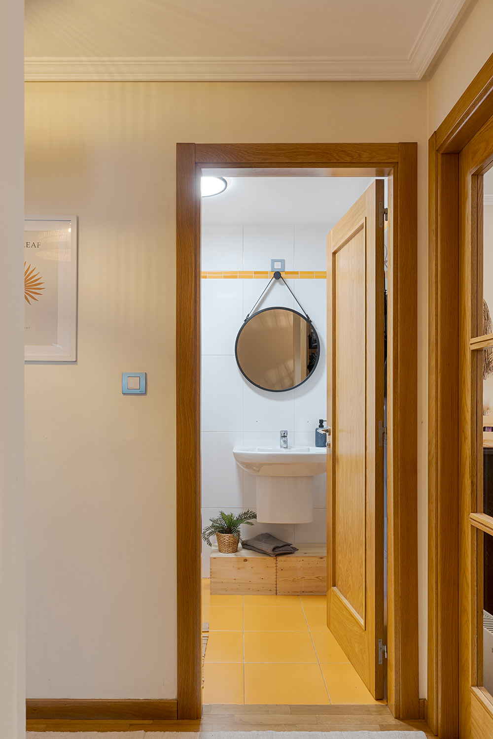 Vista desde el pasillo hacia un baño con azulejos amarillos, lavabo blanco y espejo redondo. La puerta de madera del baño está abierta, y la pared del pasillo tiene un cuadro decorativo.