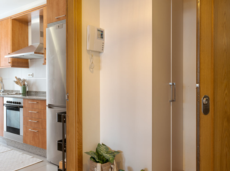 Entrada de la casa con puerta de madera abierta, vista parcial de una cocina moderna con electrodomésticos de acero inoxidable y armarios de madera. En la pared hay un intercomunicador y una planta en maceta.