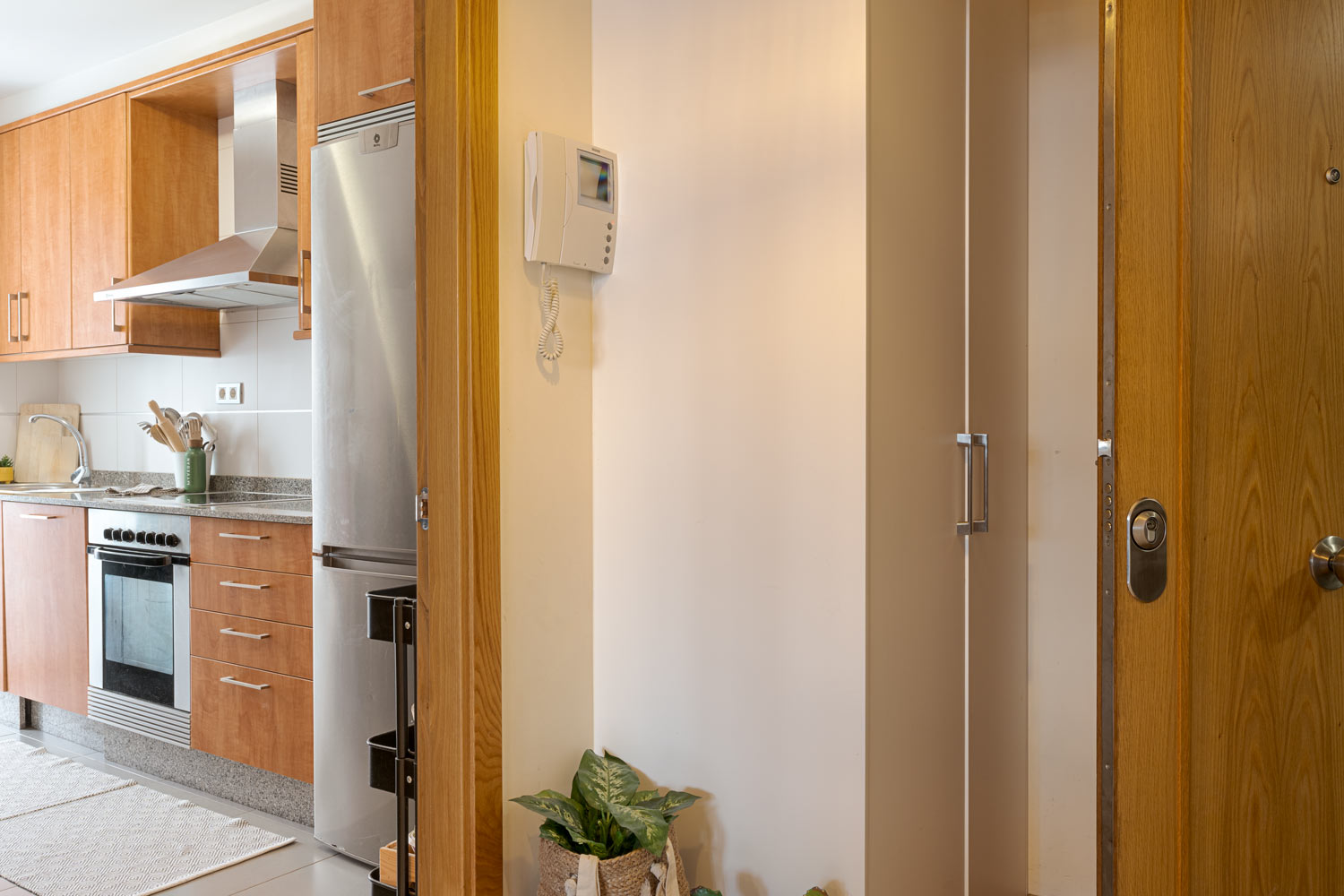 Entrada de la casa con puerta de madera abierta, vista parcial de una cocina moderna con electrodomésticos de acero inoxidable y armarios de madera. En la pared hay un intercomunicador y una planta en maceta.