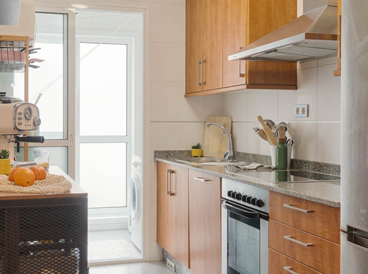 Cocina moderna con armarios de madera, encimeras de granito, electrodomésticos de acero inoxidable y una isla con naranjas. La cocina tiene una puerta de vidrio que conduce a una zona de lavandería.