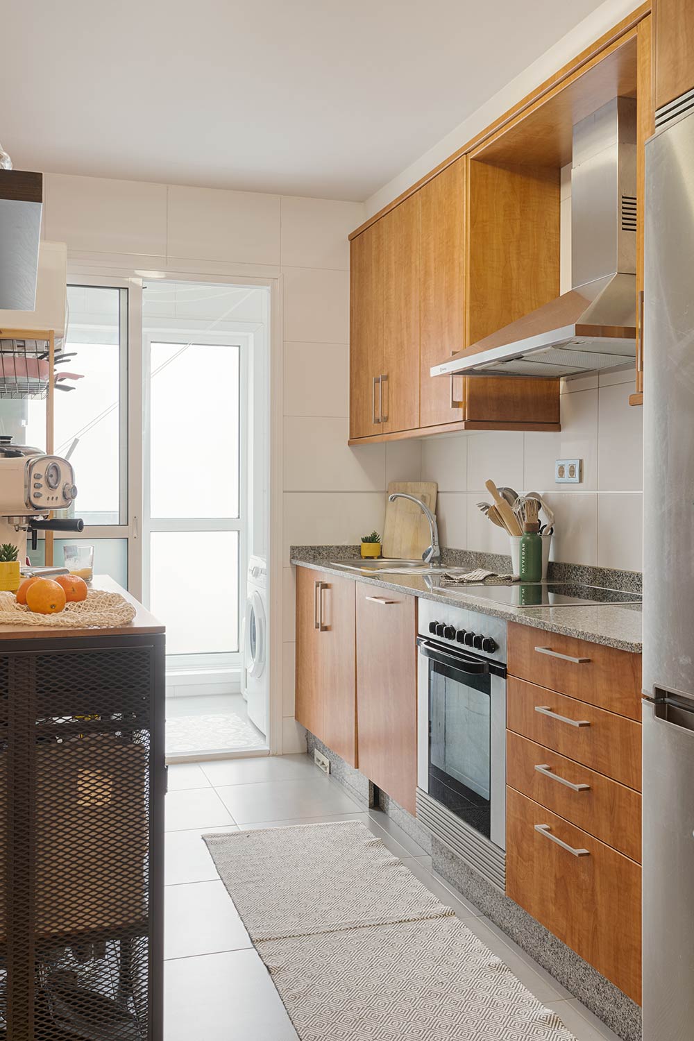 Cocina moderna con armarios de madera, encimeras de granito, electrodomésticos de acero inoxidable y una isla con naranjas. La cocina tiene una puerta de vidrio que conduce a una zona de lavandería.