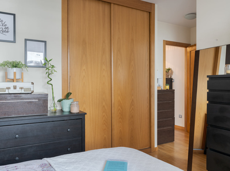 Dormitorio con armario empotrado de puertas correderas de madera, cómoda y espejo grande. La habitación está decorada con plantas, cuadros y una maleta vintage sobre la cómoda.