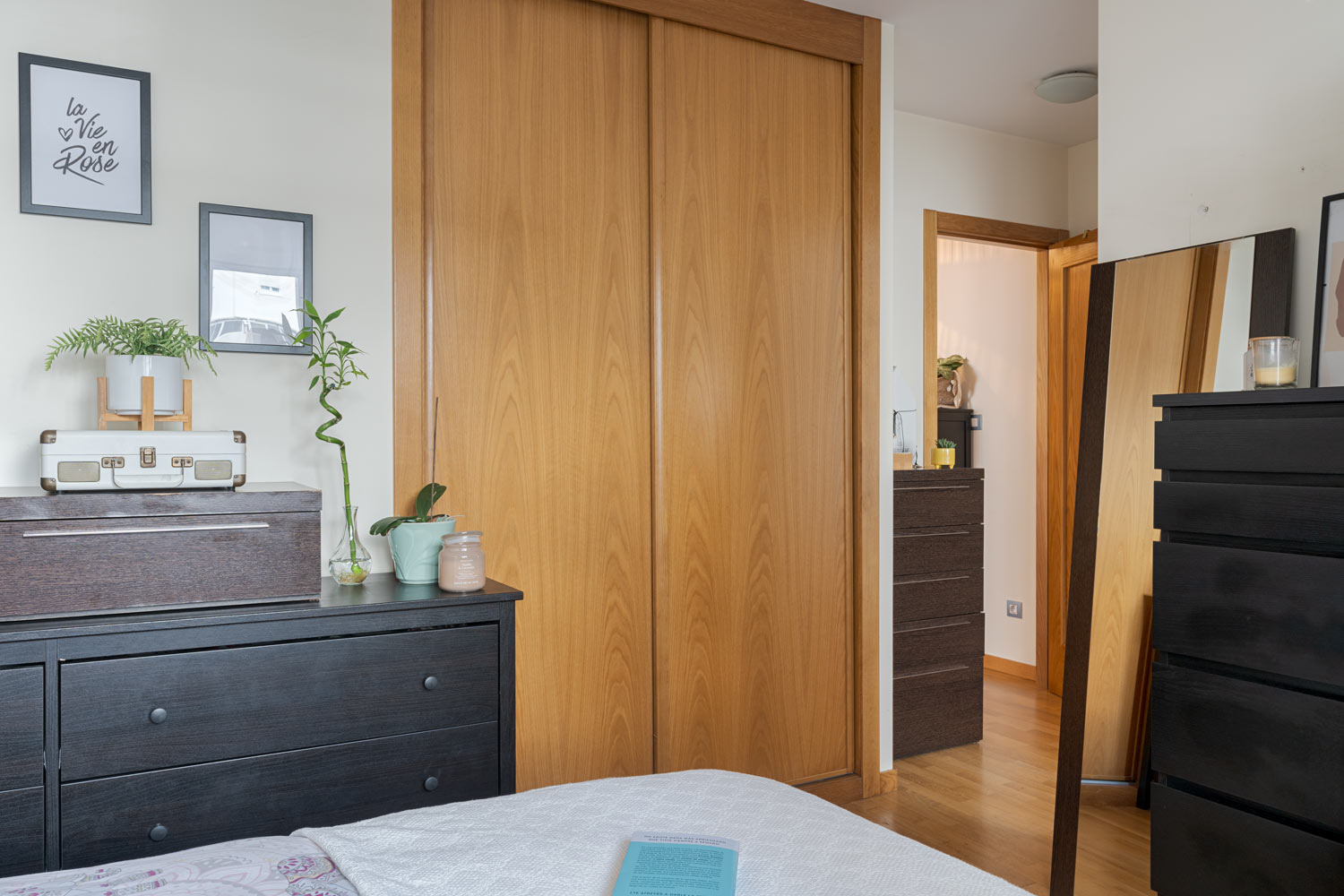 Dormitorio con armario empotrado de puertas correderas de madera, cómoda y espejo grande. La habitación está decorada con plantas, cuadros y una maleta vintage sobre la cómoda.