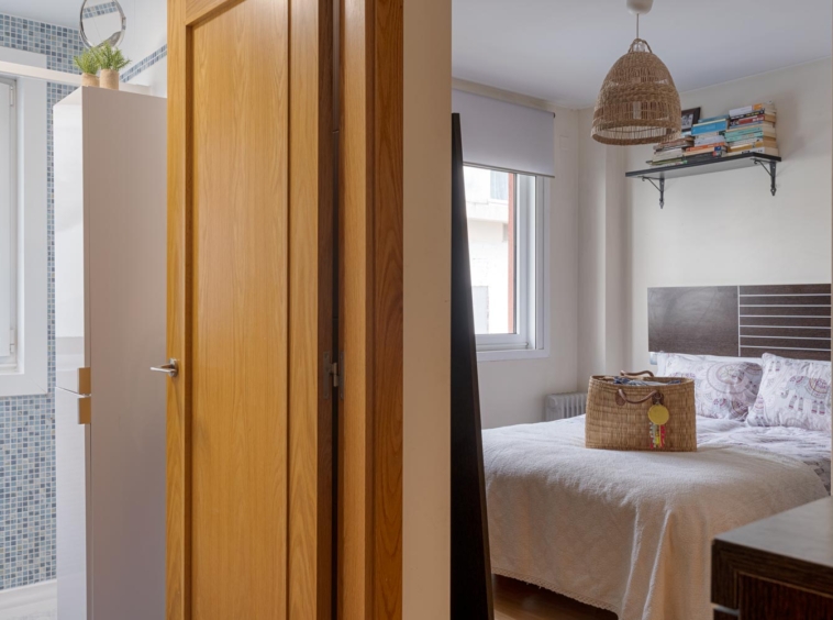 Vista desde el pasillo de un dormitorio con cama doble, ventana grande, lámpara de mimbre colgante y estantería con libros. A la derecha se ve una puerta que conduce a un baño con azulejos azules.