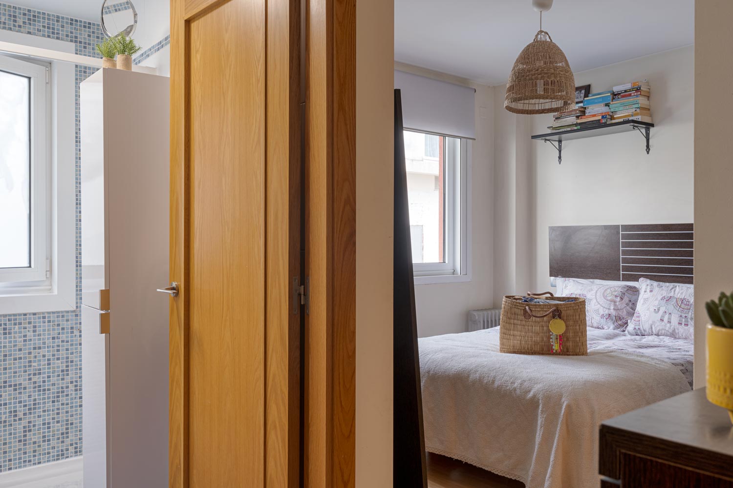 Vista desde el pasillo de un dormitorio con cama doble, ventana grande, lámpara de mimbre colgante y estantería con libros. A la derecha se ve una puerta que conduce a un baño con azulejos azules.