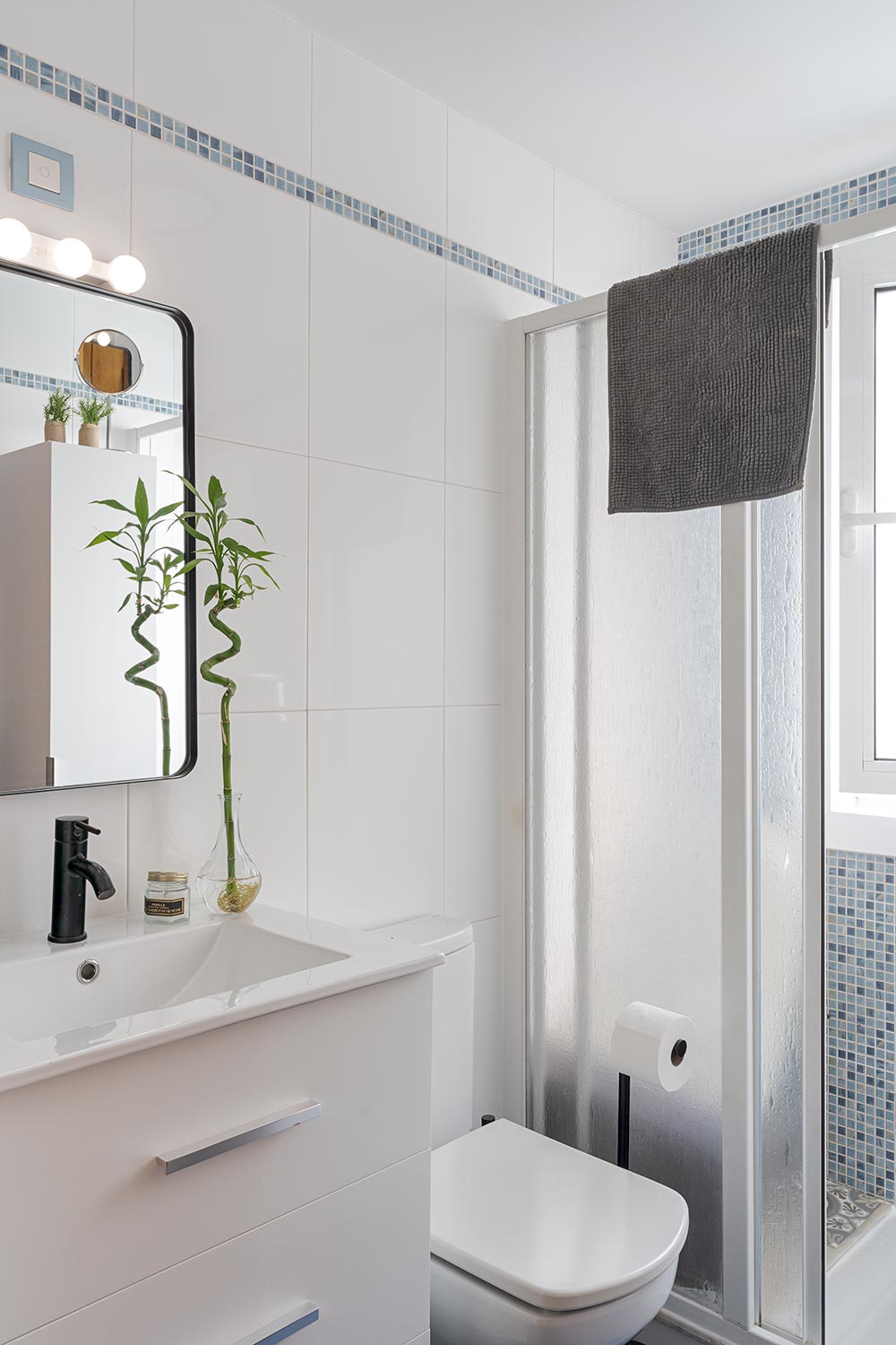 Baño moderno con lavabo blanco, espejo grande con luces, inodoro y ducha con mampara de vidrio esmerilado. El baño está decorado con plantas y azulejos en tonos azules.