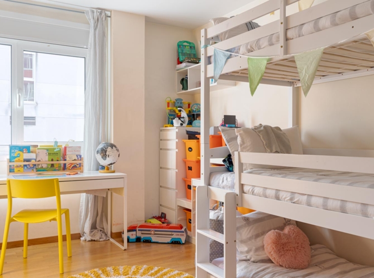 Habitación infantil con litera blanca, escritorio con silla amarilla, estantería con juguetes y decoración colorida. La habitación tiene una ventana grande que permite la entrada de luz natural.