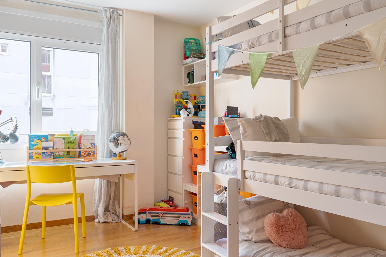 Habitación infantil con litera blanca, escritorio con silla amarilla, estantería con juguetes y decoración colorida. La habitación tiene una ventana grande que permite la entrada de luz natural.