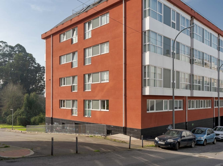 Fachada de un edificio residencial de color rojo con amplias ventanas y varios coches estacionados en la calle. El entorno incluye áreas verdes y árboles.