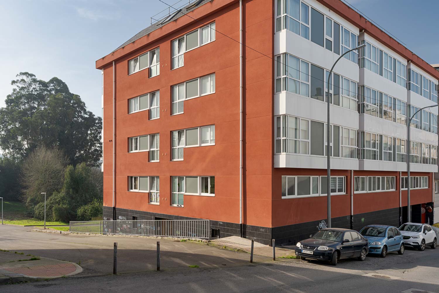 Fachada de un edificio residencial de color rojo con amplias ventanas y varios coches estacionados en la calle. El entorno incluye áreas verdes y árboles.