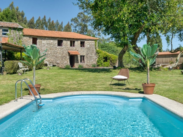 Casa rústica en venta en Aranga por Inmobiliaria Morando, con piscina, fachada de piedra, tejado de tejas rojas y jardín amplio con árboles.