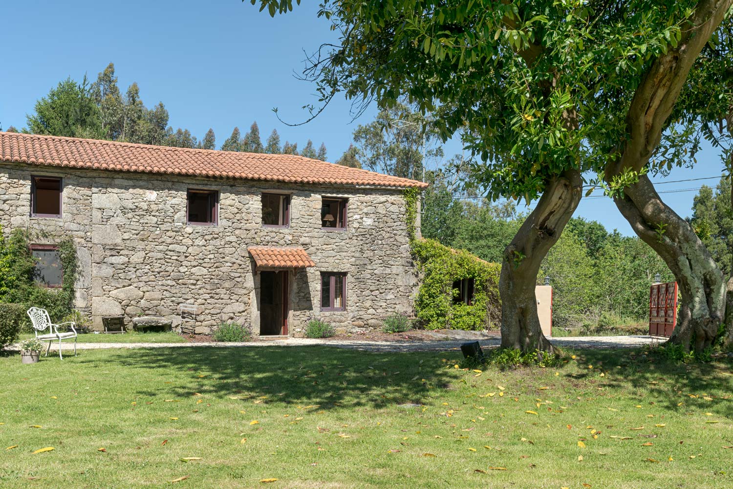 Casa rústica en venta en Aranga por Inmobiliaria Morando, con fachada de piedra, tejado de tejas rojas, y jardín amplio con árboles.