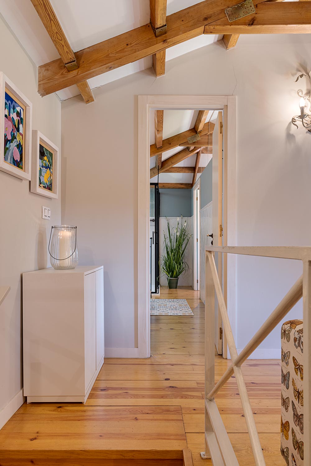 Vista del pasillo con vigas de madera en el techo y piso de madera, que conecta a un baño con plantas decorativas en la distancia. Casa en venta por Morando en Aranga.