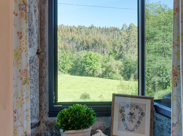 Vista desde la ventana de una casa en venta en Aranga por Morando, mostrando un paisaje verde y un pequeño escritorio azul con plantas y decoración.