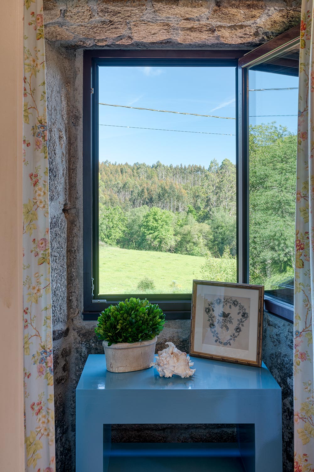 Vista desde la ventana de una casa en venta en Aranga por Morando, mostrando un paisaje verde y un pequeño escritorio azul con plantas y decoración.