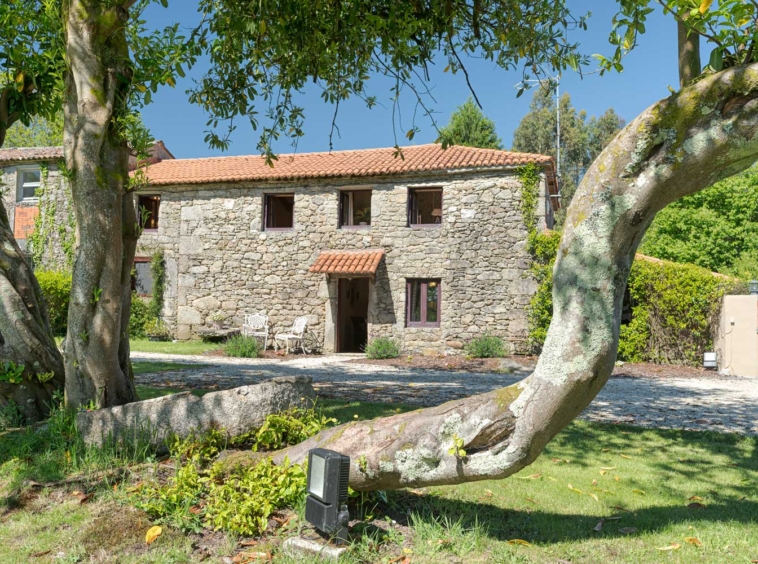 Casa rústica en venta en Aranga por Inmobiliaria Morando, con fachada de piedra, tejado de tejas rojas, jardín amplio con árboles y muebles de jardín.