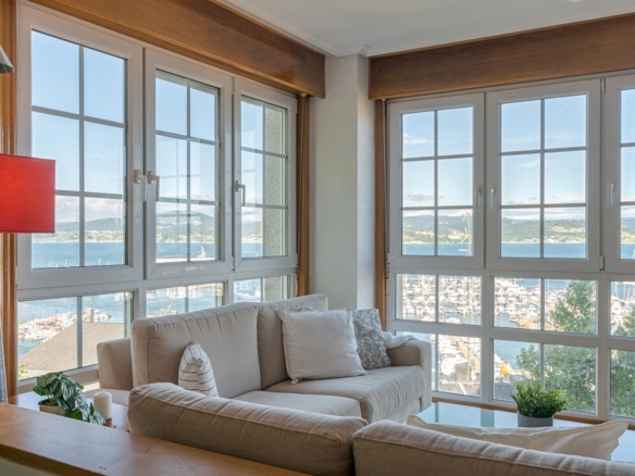 "Vista de un salón moderno con grandes ventanales que ofrecen una impresionante vista al puerto deportivo y al mar en Sada. El salón está decorado con sofás beige, cojines blancos y una lámpara de pie con pantalla roja. Se puede apreciar la luz natural que inunda el espacio y resalta la comodidad y el ambiente acogedor del lugar."